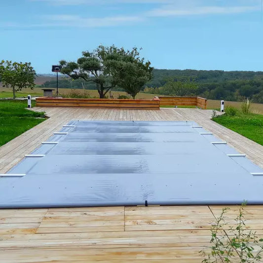 A manual Safety pool cover laid out on a wooden deck, covering a rectangular pool with a tree and sky in the background.